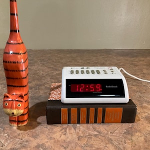 Puede incluir: Una figura alta de gato de madera tallada a mano, de color naranja con rayas negras, junto a un reloj despertador digital blanco RadioShack con pantalla LED roja que muestra las 12:59, sobre un libro marrón con detalles naranjas en el lomo.