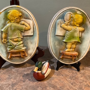 May include: Two ceramic wall plaques depicting a child in a green and yellow outfit sitting on a stool. The child on the left is drawing on a piece of paper, while the child on the right is looking in a mirror. A small wooden duck figurine is in the foreground.