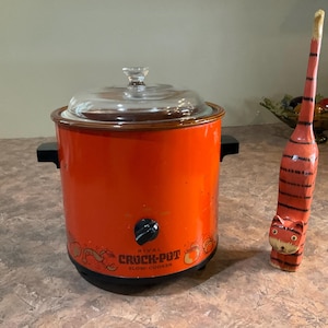 May include: An orange Rival Crock-Pot slow cooker with a glass lid and black handles. A decorative, hand-painted wooden cat figurine with red and black stripes stands to the right.
