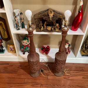 May include: Two tall, carved wooden lamps with white lampshades. The lamps are on a white shelf with various Christmas decorations, including a nativity scene, a nutcracker, and a red and green elf.