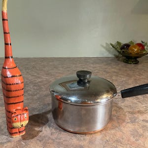 Puede incluir: Una figura de gato de madera alta de color naranja con rayas horizontales negras y una cola larga y erguida, junto a un cazo de acero inoxidable con tapa, mango negro y una pequeña anilla de metal en el extremo del mango.