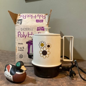 May include: A white electric coffee pot with a brown base and a floral design. The pot has a handle and a spout. The box reads "Poly Hot Pot" and "Regal".