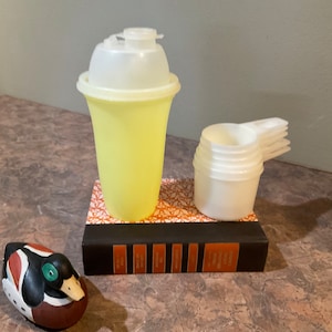 May include: A yellow plastic shaker with a white lid and a stack of four white plastic measuring cups on a book with a brown cover.