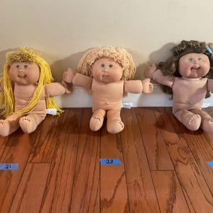 May include: Three vintage Cabbage Patch Kids dolls with different hair colours: yellow, light brown, and dark brown. The dolls are sitting on a wooden floor with their arms outstretched. Each doll has a blue tag with a number.