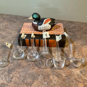 Puede incluir: Seis cucharones de vidrio transparente con mangos, numerados del 1 al 5, dispuestos sobre un libro con un pato pintado en la parte superior.