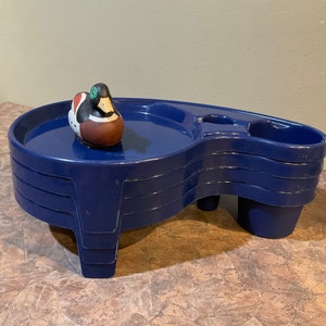 May include: A set of four blue plastic serving trays with a duck figurine on top. The trays are stacked and have a raised edge and a handle on one side. The trays are designed to be used as a serving stand.