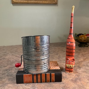 May include: A vintage metal sifter with a red handle and the text "GUARANTEED". Beside it is a carved wooden cat figurine with orange and black stripes. The objects rest on a stack of books.