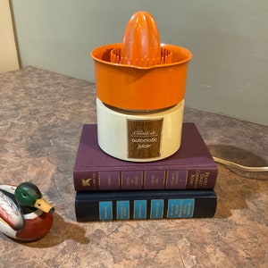 May include: An orange and white electric juicer with a wooden label that reads "automatic juicer". The juicer is sitting on top of a stack of three books.