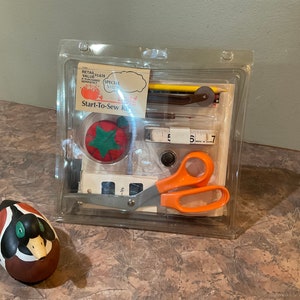 May include: A clear plastic package containing a Start-To-Sew Kit with sewing supplies, including scissors, needles, thread, a thimble, a tape measure, and a pencil. The package is on a brown countertop next to a small, painted wooden duck.