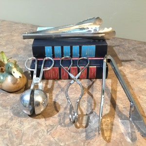 以下が含まれることがあります： 氷用トング、サラダ用トング、サービングトング、スプーンの4つの銀製のキッチン用トングのセット。トングは、セラミック製の鴨の置物と2冊の青い本のあるテーブルの上に置かれています。