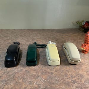 以下が含まれることがあります： 黒、ダークグリーン、クリーム、ベージュの壁掛け電話4台。それぞれ受話器とカールコードが付いています。電話の横には、オレンジと黒の縞模様が入った、背が高く細身の猫の置物が立っており、長い尻尾をまっすぐ上に伸ばしています。