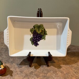 May include: White ceramic baking dish with a grape design. The dish has a raised rim and two handles. The dish is on a wooden stand.