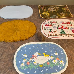 May include: A selection of decorative placemats with diverse patterns. Features a white oval mat, a yellow daisy-shaped mat, a green floral mat, a winter scene mat, and a blue mat with a duck and duckling design.