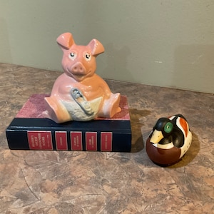 Peut inclure: Une tirelire en céramique rose avec une couche bleue et une épingle de sûreté, posée sur un livre bleu et rouge. Un canard en céramique brun et vert est posé sur une surface brune.