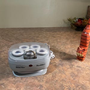 May include: A gray electric hair setter with a clear plastic lid containing five white rollers and a metal wire clip, alongside a tall, slender, orange and black striped wooden cat figurine with a long tail and painted facial features.