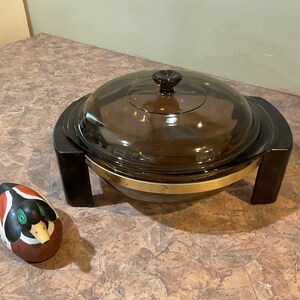 May include: A brown glass casserole dish with a lid and a gold rim. The dish is sitting on a black stand with two legs. A small wooden duck figurine is in the foreground.