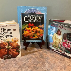 May include: Three cookbooks on a table. The first cookbook is titled "All-Time Favorite Recipes" by Better Homes and Gardens. The second cookbook is titled "Treasury of Country Recipes" by Land O' Lakes. The third cookbook is titled "Martha Stewart's Hors d'Oeuvres".