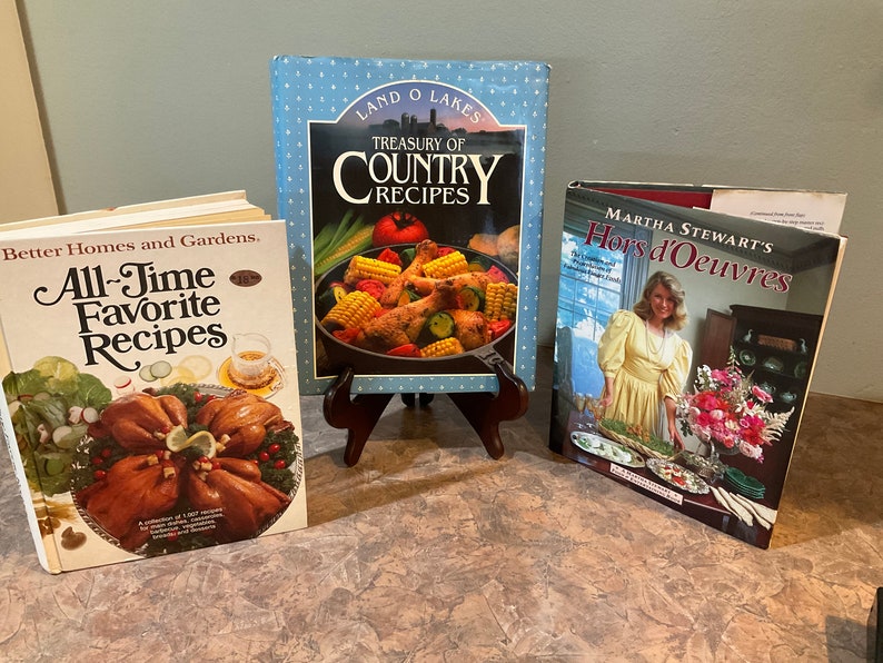 May include: Three cookbooks on a table. The first cookbook is titled "All-Time Favorite Recipes" by Better Homes and Gardens. The second cookbook is titled "Treasury of Country Recipes" by Land O' Lakes. The third cookbook is titled "Martha Stewart's Hors d'Oeuvres".