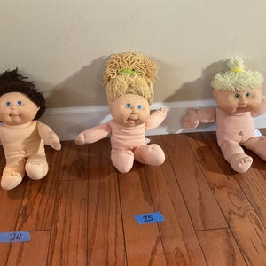 Pode incluir: Três bonecas Cabbage Patch Kids vintage com diferentes penteados. As bonecas têm olhos azuis e usam roupas rosa claro. Cada boneca está sentada em um chão de madeira. Há etiquetas azuis com números no chão.