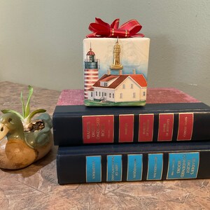 May include: A stack of three hardback books with red, blue and turquoise spines. The top book has a red spine with gold lettering that reads "Reader's Digest Condensed Books". The middle book has a blue spine with gold lettering that reads "The Indian" and "Sinnequin". The bottom book has a turquoise spine with gold lettering that reads "Lady of the Island". A small, white gift box with a red bow and a lighthouse illustration sits on top of the books. A ceramic duck planter with a green glaze and a small succulent plant sits on the left side of the image.