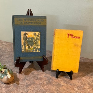May include: Two vintage hardcover books on display stands. The book on the left has a blue cover with the title "The Wonderland of Knowledge" and a picture of three people standing around a globe. The book on the right has a yellow cover with the title "Tail Waggers" in red lettering.