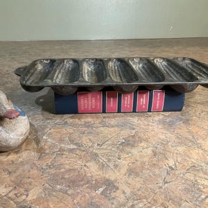 May include: A cast iron cornbread pan shaped like ears of corn, resting on a blue book with red spines. A decorative duck figurine with blue and white paint and a pink beak is to the left.