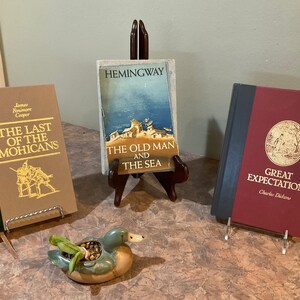 Op de afbeelding: Drie hardcover boeken op presentatiestandaards. De boeken zijn "The Last of the Mohicans" van James Fenimore Cooper, "The Old Man and the Sea" van Hemingway en "Great Expectations" van Charles Dickens. Op de voorgrond staat een kleine keramische eendenpot met een groene en bruine glazuur.