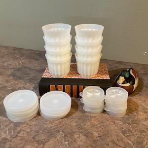 May include: A set of white plastic cups, some stacked and some in a pile, with a duck decoy and a book. The cups have a fluted design and are ideal for making individual desserts.