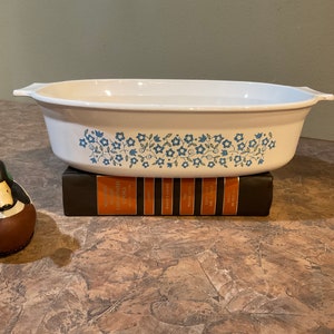 Puede incluir: Plato para hornear ovalado blanco con un patrón floral azul. El plato está sobre una pila de libros.