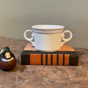 Op de afbeelding: Een witte keramische soepkom met twee handvatten, rustend op een decoratief boek. Een beschilderd eendenbeeldje met bruine, zwarte en groene details staat links. De kom heeft een lichtgrijze band.