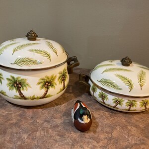 Może przedstawiać: Dwa białe ceramiczne naczynia do zapiekania z pokrywkami, zdobione palmami i paprociami. Naczynia mają uchwyty i gałki w kolorze brązu. Obecna jest również mała, dekoracyjna figurka kaczki.