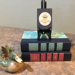 Puede incluir: Dos libros de tapa dura azules con letras doradas en los lomos. El libro superior tiene una cubierta de mármol verde y blanco. El libro inferior tiene una cubierta de mármol rojo y blanco. Un pequeño soporte de madera con un sello negro y dorado está delante de los libros. Un macetero de pato de cerámica con una planta suculenta está en primer plano.