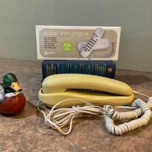 May include: A yellow corded rotary phone with a coiled cord. The phone is sitting on a stack of books. The box for a "super trim phone" is behind the books. A painted wooden duck figurine is in the foreground.