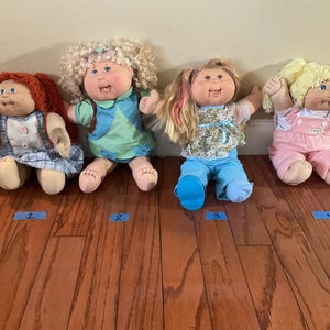 Puede incluir: Cuatro muñecas vintage Cabbage Patch Kids se exhiben sobre un suelo de madera. Cada muñeca tiene un peinado y un atuendo únicos, que incluyen vestidos, monos y tops coloridos. Las muñecas están numeradas del 1 al 4 con cinta azul.