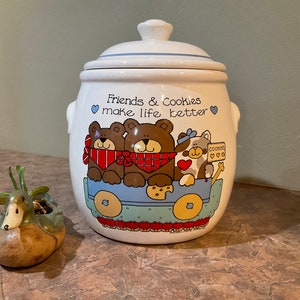 Puede incluir: Jarra de galletas de cerámica blanca con tapa azul y una imagen de dibujos animados de tres osos y un gato montando en un carro. La jarra tiene el texto "Friends & Cookies make life better" en la parte delantera.