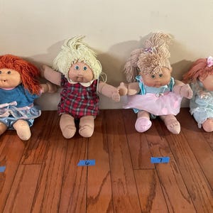 Può includere: Quattro bambole vintage Cabbage Patch Kids sono esposte su un pavimento in legno. Ogni bambola ha un'acconciatura e un abito unici, tra cui vestiti e fiocchi. Le bambole sono numerate con nastro adesivo blu.
