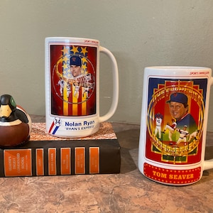 May include: Two white ceramic mugs with baseball player images. The mug on the left features Nolan Ryan with the text "Nolan Ryan" and "Ryan's Express". The mug on the right features Tom Seaver with the text "Tom Seaver".