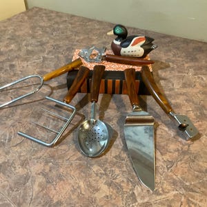 May include: A set of five kitchen utensils with brown plastic handles. The utensils include a fork, a slotted spoon, a spatula, a cheese grater, and a can opener. The utensils are arranged on a wooden surface with a decorative duck figurine.