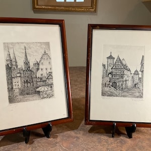 May include: Two framed black and white etchings depicting European city scenes with historic architecture, including a fountain and timber-framed buildings. Each print is housed in a dark brown wooden frame with a white mat and glass front.