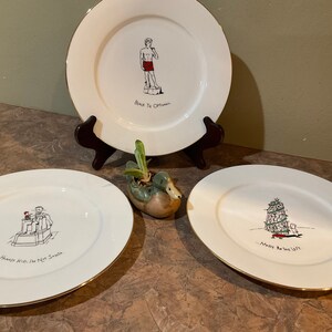 May include: Three white ceramic plates with gold trim and humorous illustrations. The plates feature a man in a red bathing suit, a man in a Santa hat, and a Christmas tree.
