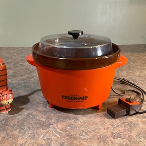 May include: A bright orange Rival Crock-Pot slow cooker with a brown ceramic insert, a clear glass lid, and a brown power cord with a control dial. Beside it stands a tall, slender, orange and black striped wooden cat figurine with a long tail.