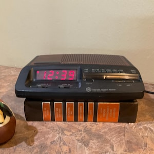 Puede incluir: Un radio despertador digital negro con una pantalla LED roja que muestra la hora 12:39. El radio despertador tiene una perilla de sintonización plateada y un cable de alimentación negro.