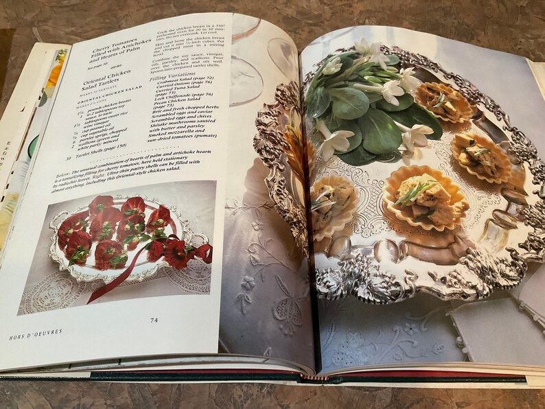 May include: An open cookbook with two pages showing recipes for Cherry Tomatoes Filled with Artichoke and Hearts of Palm and Oriental Chicken Salad Tartlets. The left page shows a silver platter with red and green tartlets. The right page shows a silver platter with white flowers and four small tartlets filled with a green and white mixture.
