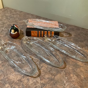 May include: Four clear glass serving dishes with a wavy design. The dishes are oval shaped and have a slight curve to them. The dishes are arranged on a brown surface.