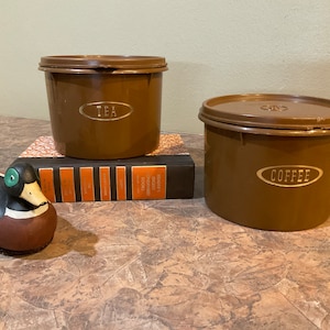Puede incluir: Dos botes de plástico marrón con tapas, uno etiquetado como "TEA" y el otro como "COFFEE". Los botes están sobre una pila de libros.