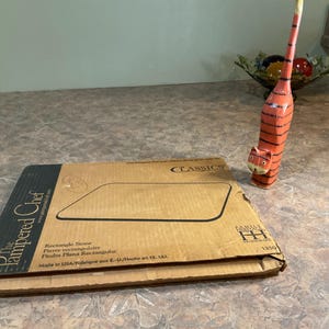 以下が含まれることがあります： The Pampered Chefの茶色の段ボール箱で、「Rectangle Stone」と表示されています。箱には黒い文字と石の黒い輪郭があります。オレンジと黒の装飾的な猫の置物が近くに立っています。