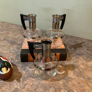 May include: Three clear glass pitchers with silver bands and black handles. The pitchers are sitting on a book with a brown cover. A small wooden duck figurine is in the foreground.