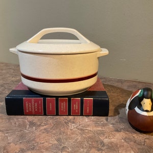 May include: A white ceramic casserole dish with a brown stripe around the top. The dish is sitting on a stack of books with the title "Reader's Digest Condensed Books". A small brown and green ceramic duck figurine is sitting on the table next to the dish.