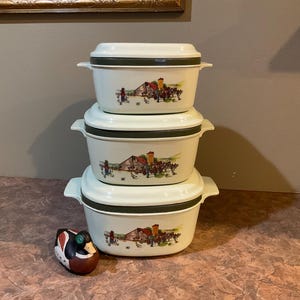 Könnte beinhalten: Ein Satz aus drei weißen Keramik-Auflaufformen mit grünem Rand und einem Landhaus-Motiv. Die Formen sind übereinander gestapelt.