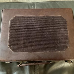 May include: A vintage brown leather briefcase with a dark brown, textured, rectangular insert on the top. The briefcase has a handle and metal clasps. The case is sitting on a dark green surface.